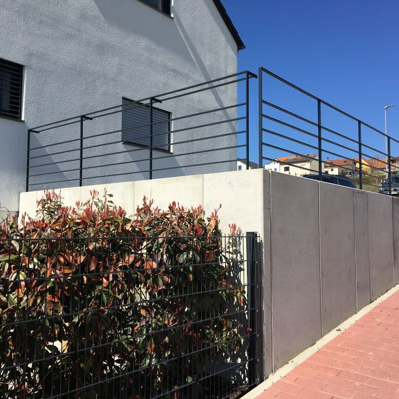Modernes Hausäußeres mit weißer Wand, schwarzem Metallgeländer und grüner Hecke unter blauem Himmel