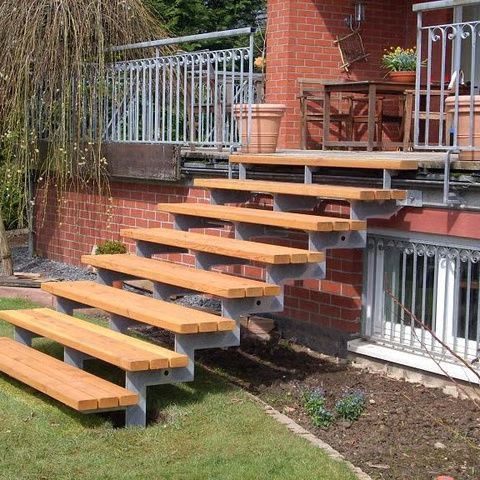 Eine hölzerne Außentreppe mit Metallstützen führt zu einer gemauerten Terrasse mit Geländer neben einem Haus.