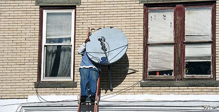 Une personne sur une échelle installe une antenne parabolique sur un bâtiment en briques, près de deux fenêtres.