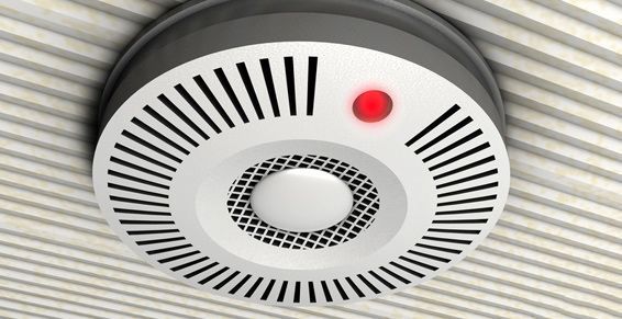 Détecteur de fumée fixé sur un plafond blanc à lattes, avec un voyant rouge allumé.