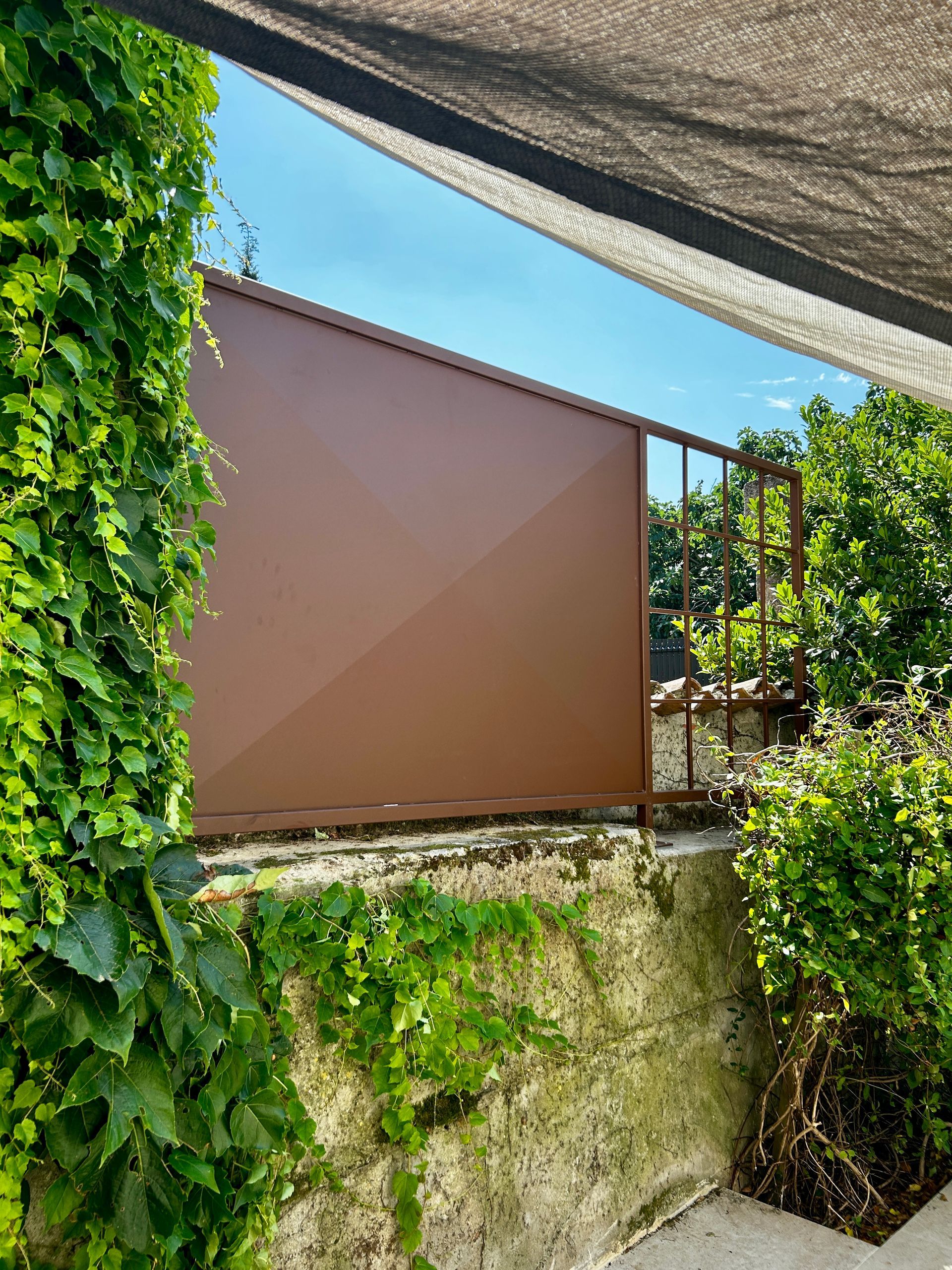 Panneau métallique brun et clôture avec plantes vertes feuillues, ciel bleu visible.