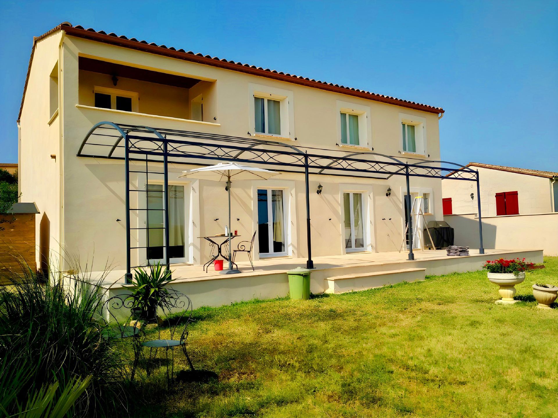 Maison à deux étages avec volets bleus, une pergola et un jardin gazonné sous un ciel bleu.