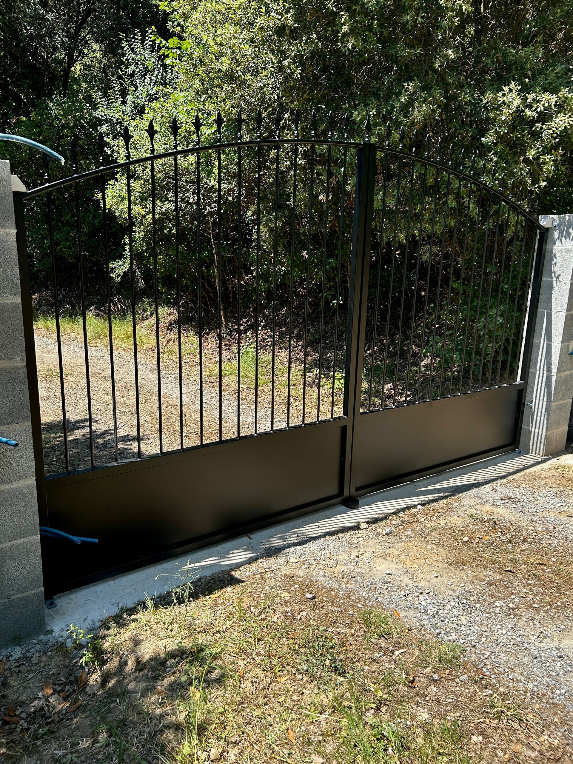 Un portail en métal noir, encadré de piliers en béton, s'ouvre sur un chemin de terre bordé d'arbres.