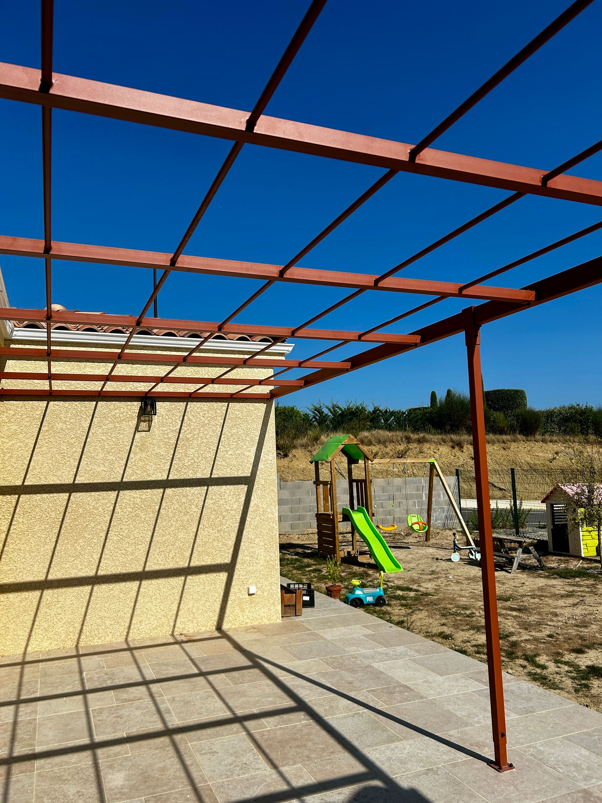 Maison beige avec terasse, une pergola effet rouillé et un ciel bleu clair.