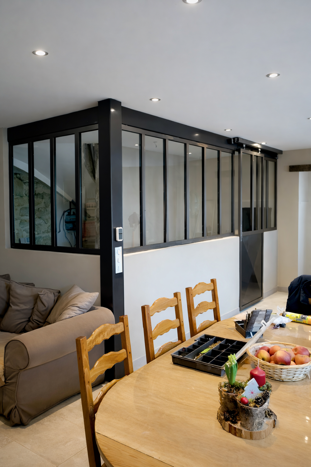 Intérieur avec table à manger, canapé et cloison vitrée. Des chaises en bois entourent la table.