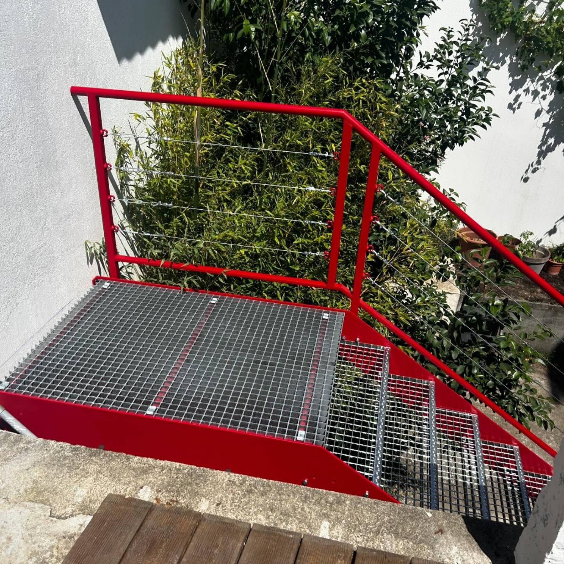 Escalier métallique rouge avec marches et rampe en caillebotis, menant à une plateforme. Feuillage vert en arrière-plan.