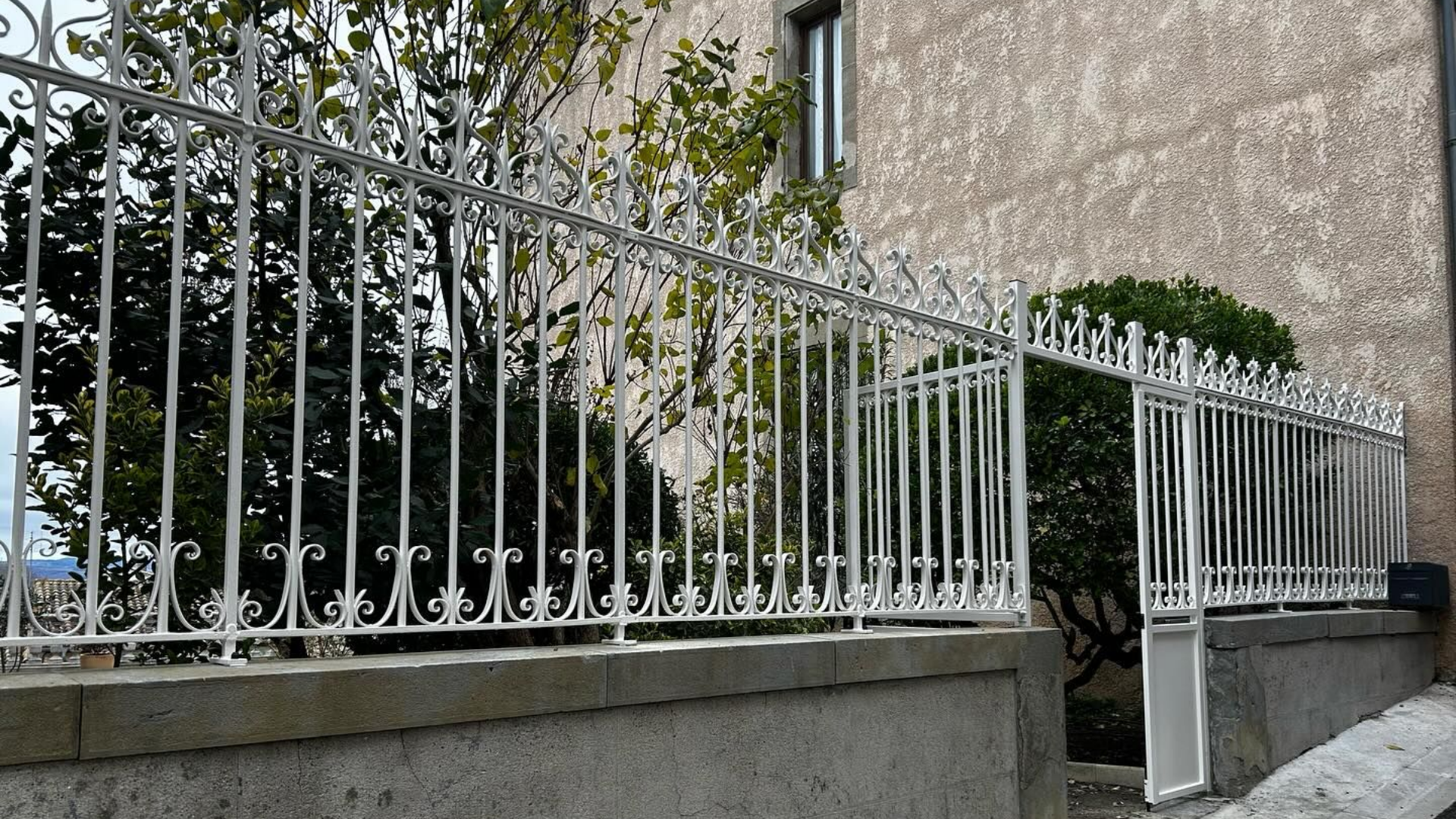 Clôture décorative en métal blanc sur un mur en béton, à côté d'un bâtiment et de verdure.