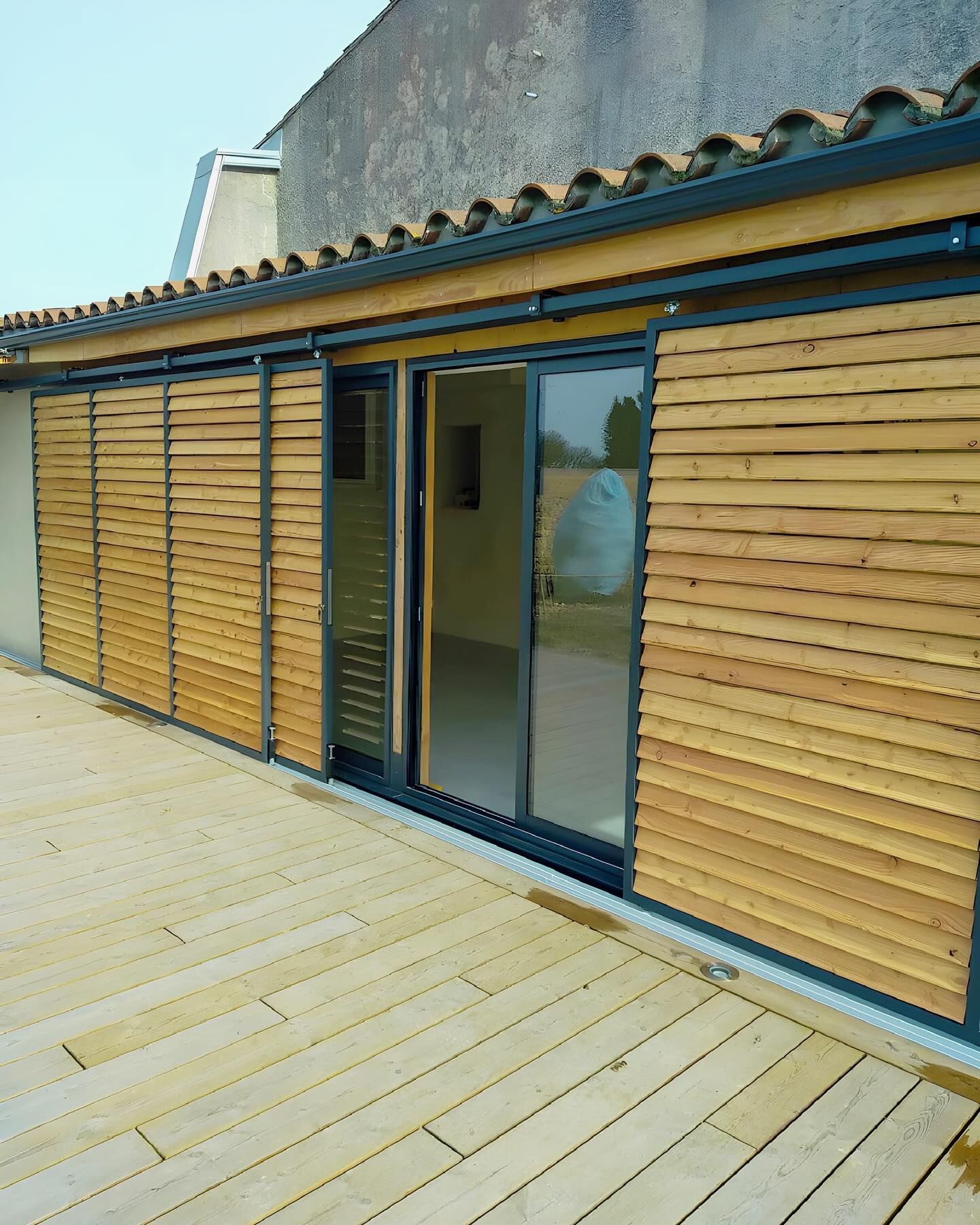 Volets en bois sur une terrasse, à côté d'une porte vitrée. Bâtiment avec un toit de tuiles.