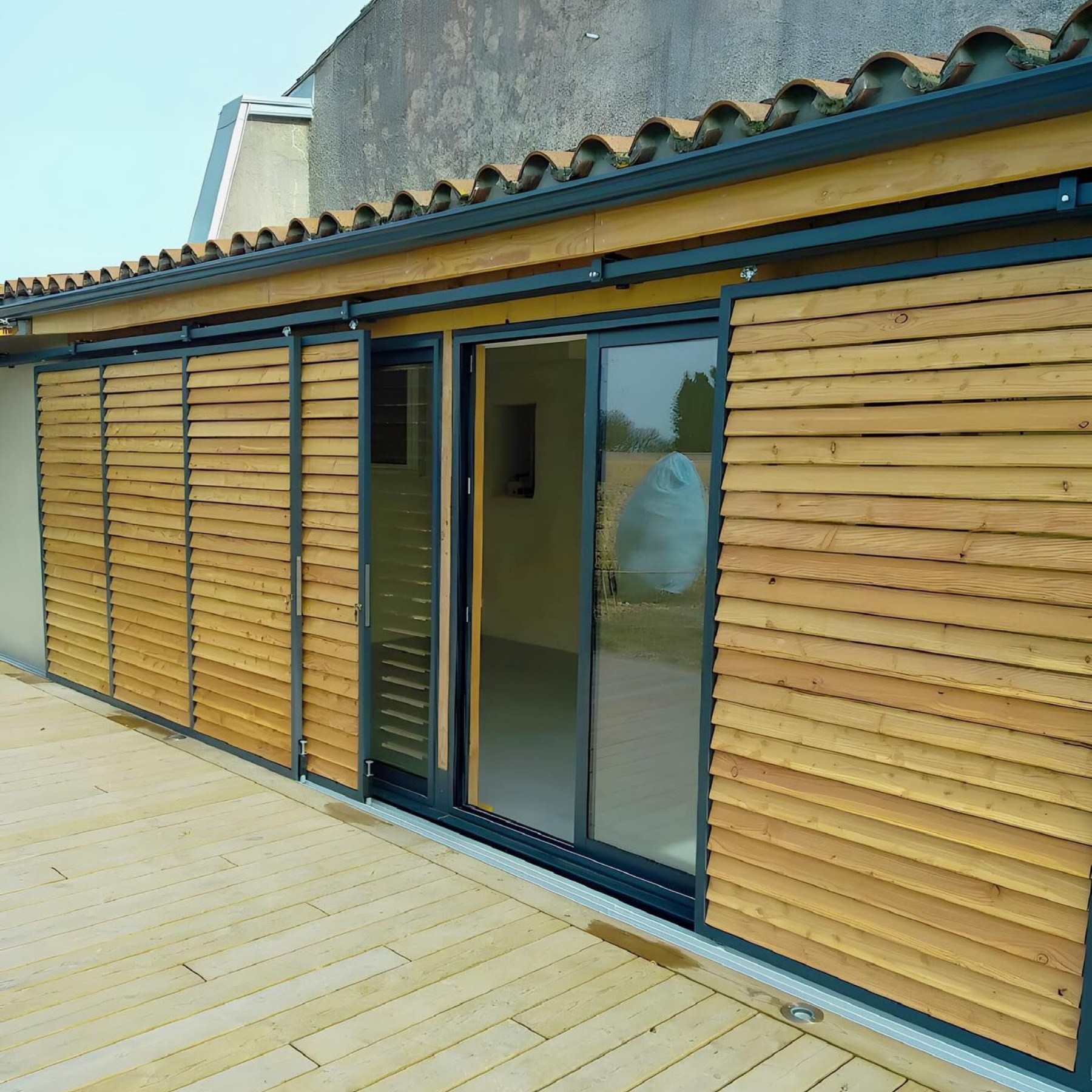 Extérieur d'un bâtiment avec volets coulissants en bois et porte vitrée. Terrasse en bois, ciel bleu.