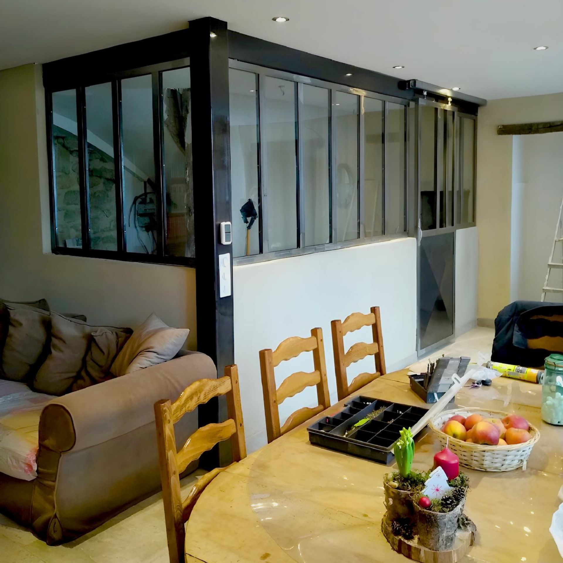 Intérieur avec table à manger, canapé et cloison vitrée. Des chaises en bois entourent la table.