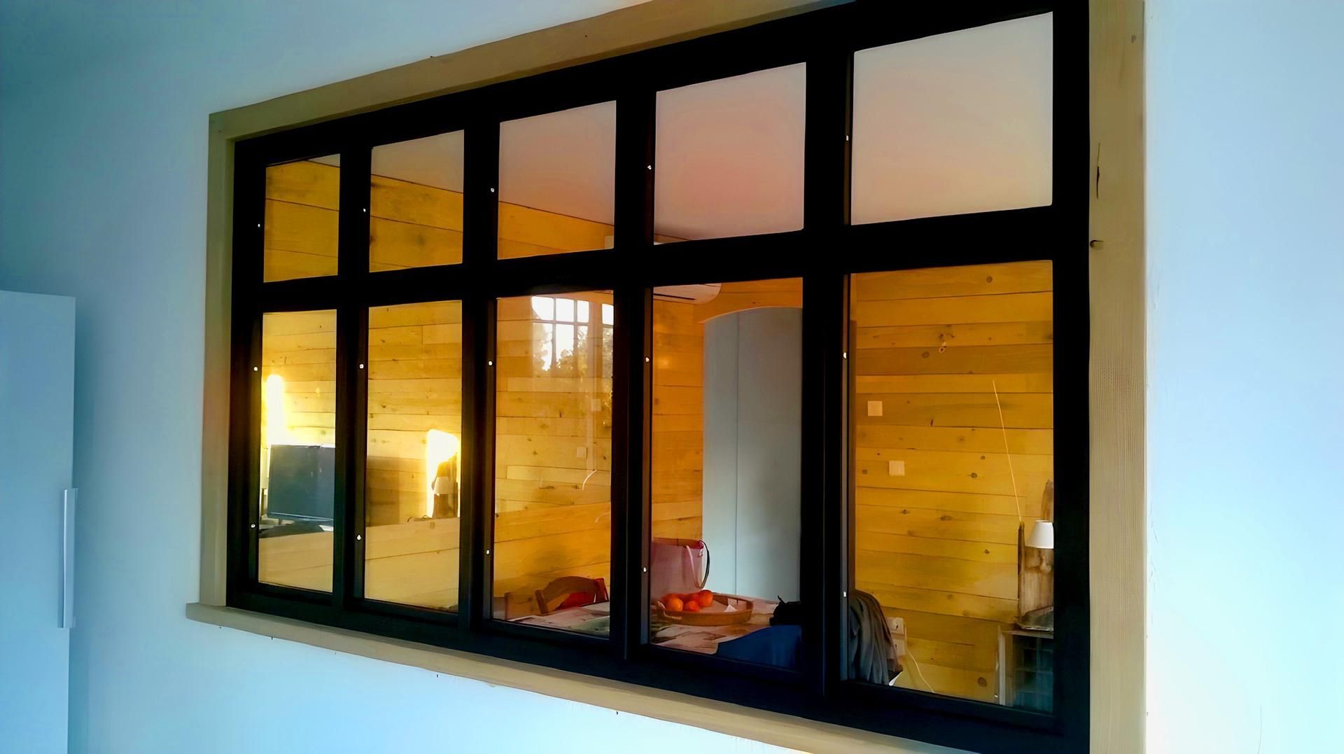 Fenêtre à plusieurs carreaux à cadre noir reflétant un intérieur en bois ; moulures beiges au mur.