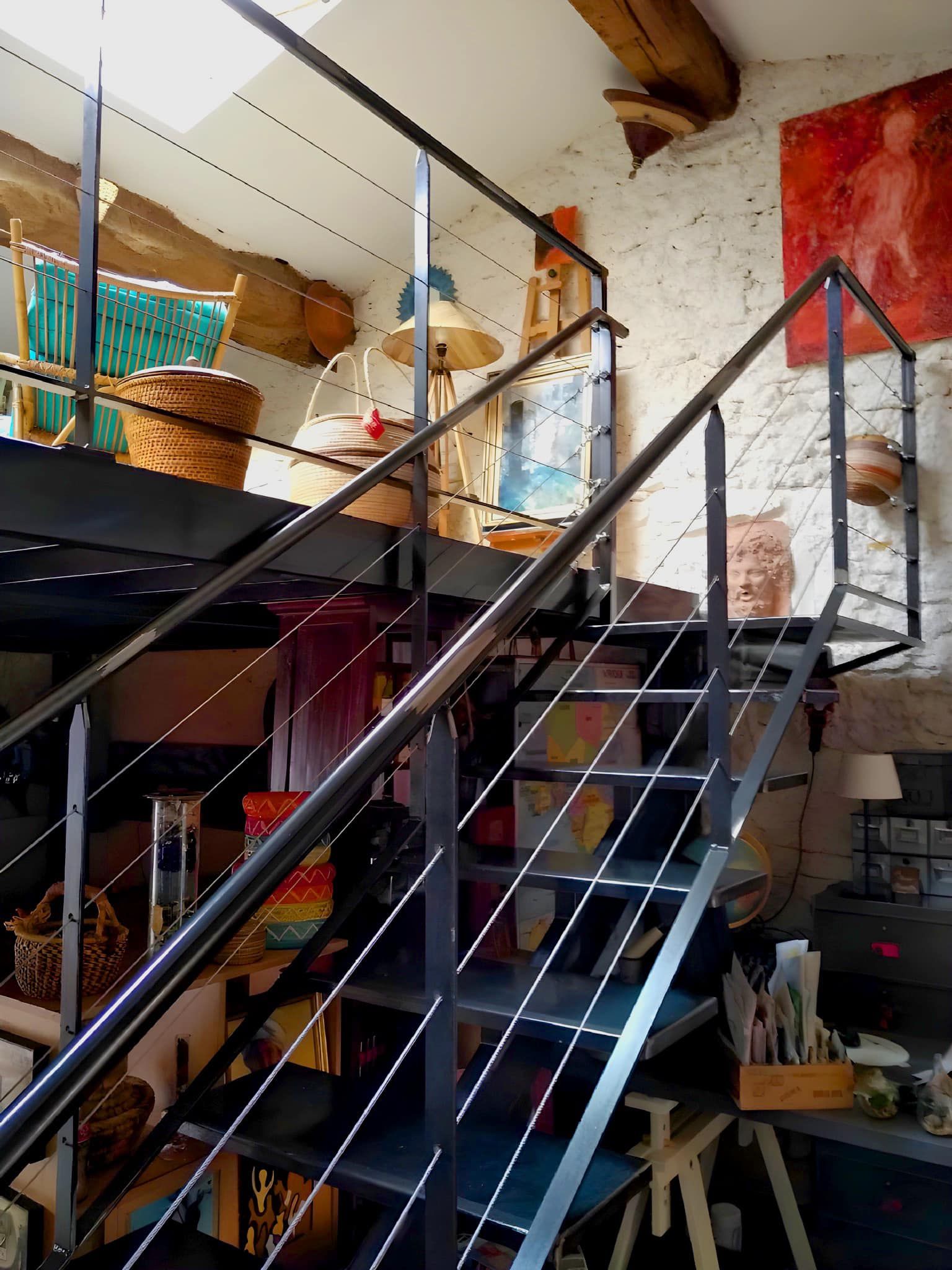 Un escalier métallique mène à une mezzanine. On y aperçoit des paniers, des œuvres d'art et des murs en briques apparentes.