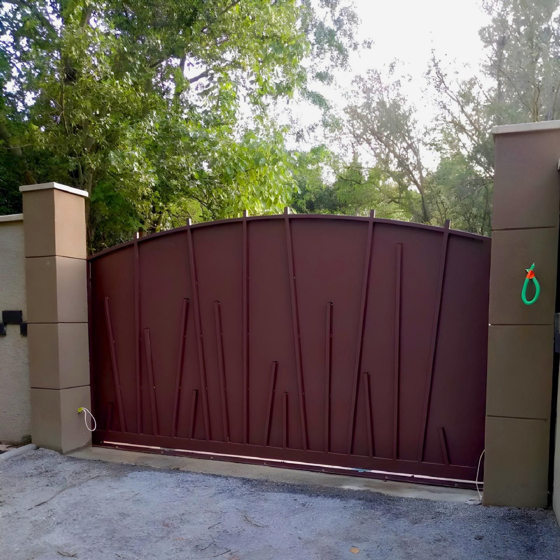 Portail en métal brun orné de lignes verticales abstraites, placé entre des piliers en béton beige, devant des arbres.