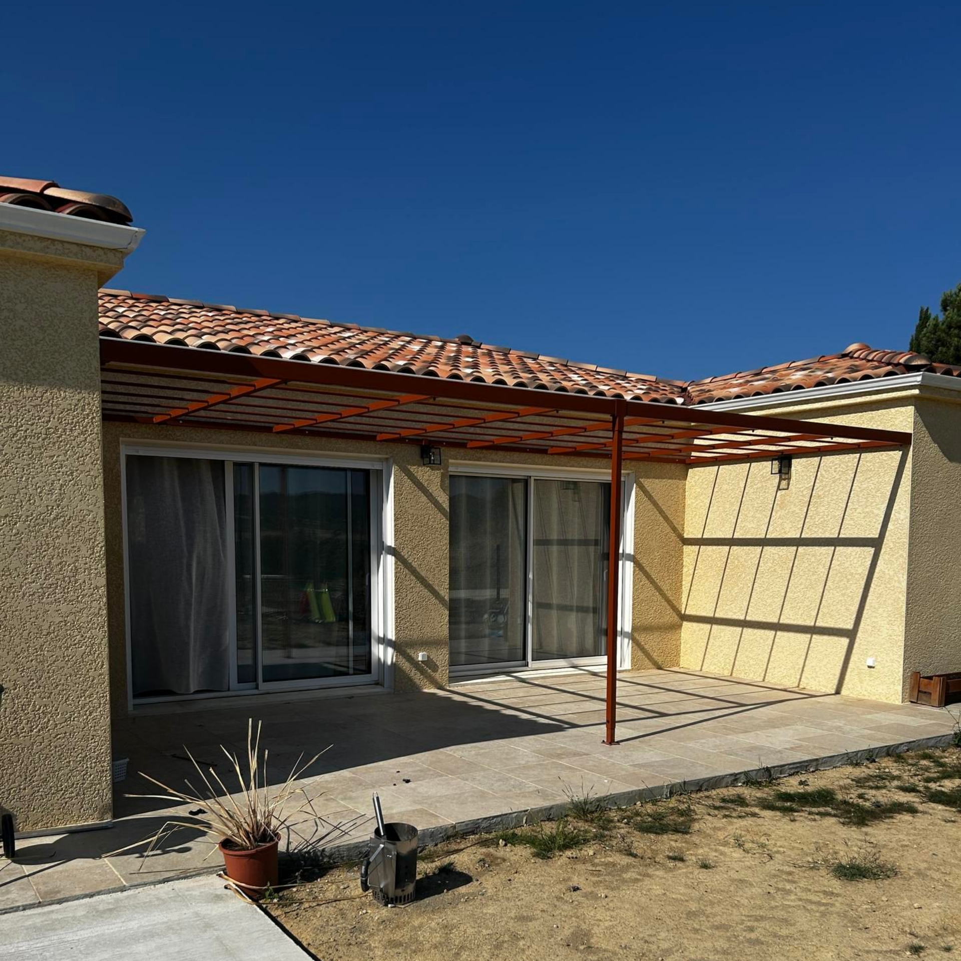 Vue extérieure d'une maison avec un toit de tuiles, une terrasse et des portes coulissantes en verre sous un ciel bleu.