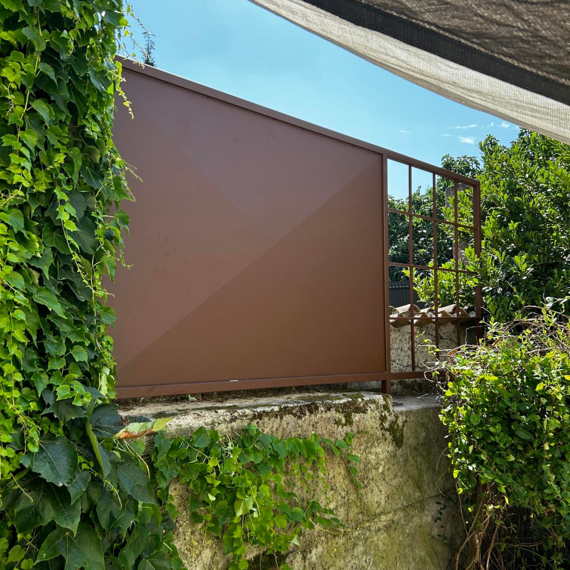 Portail et balustrade en métal brun au sommet d'un mur de pierre, recouverts d'un lierre et d'un feuillage d'un vert luxuriant.