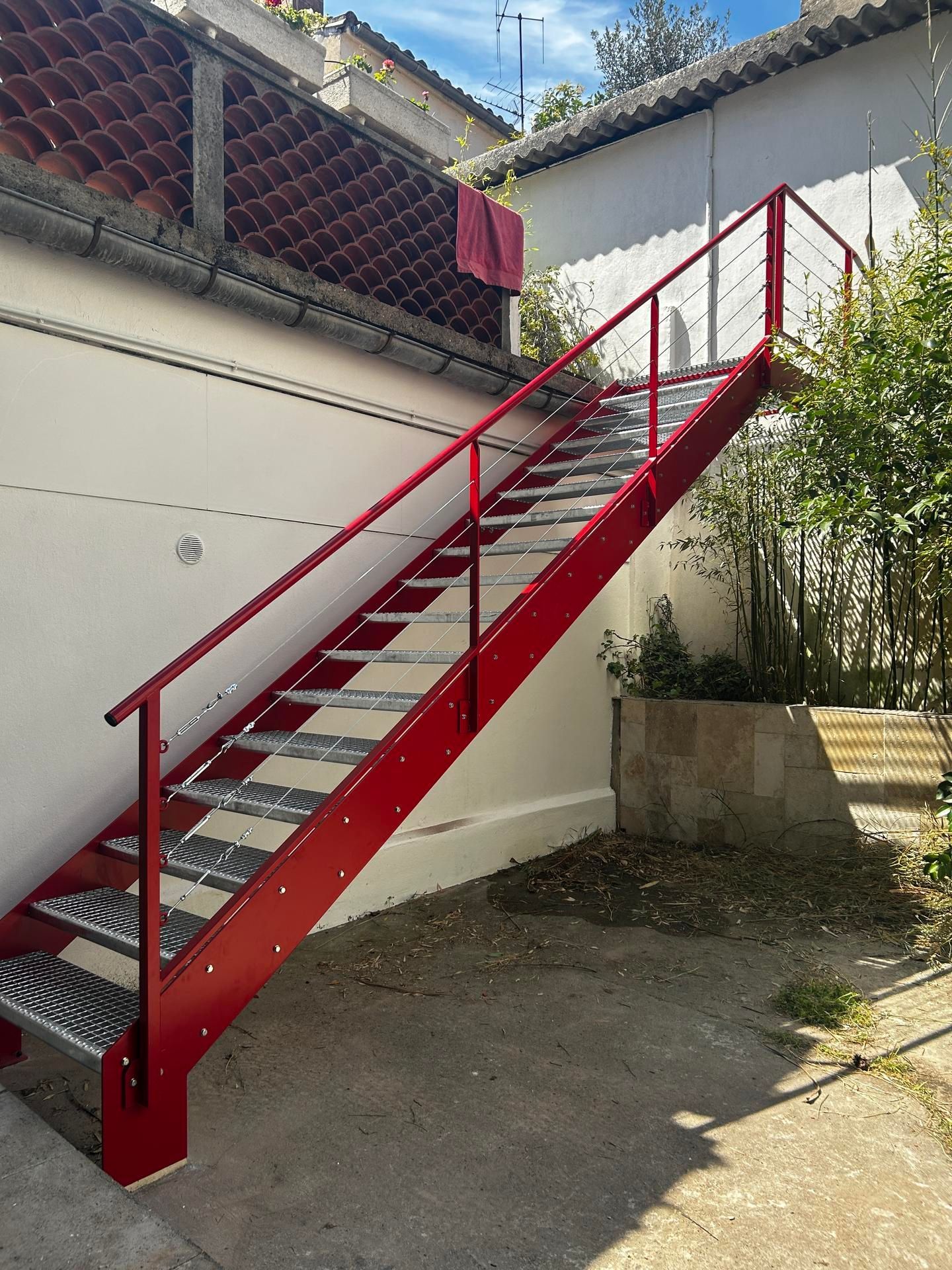 Escalier en métal rouge contre un mur blanc, avec une rambarde en câbles, menant vers l'étage.
