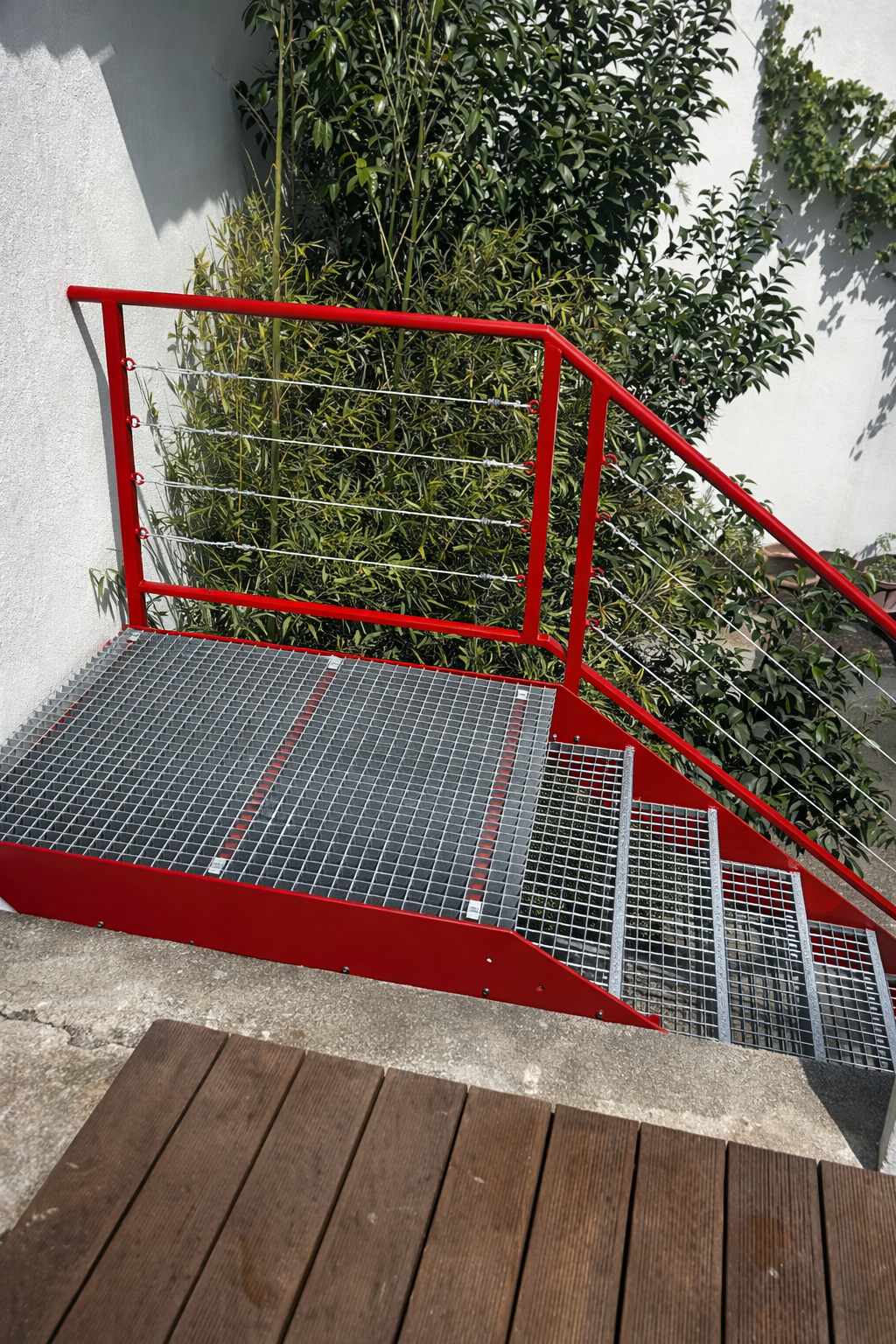 Escalier métallique rouge avec marches et rampe en caillebotis, menant à une plateforme. Feuillage vert en arrière-plan.