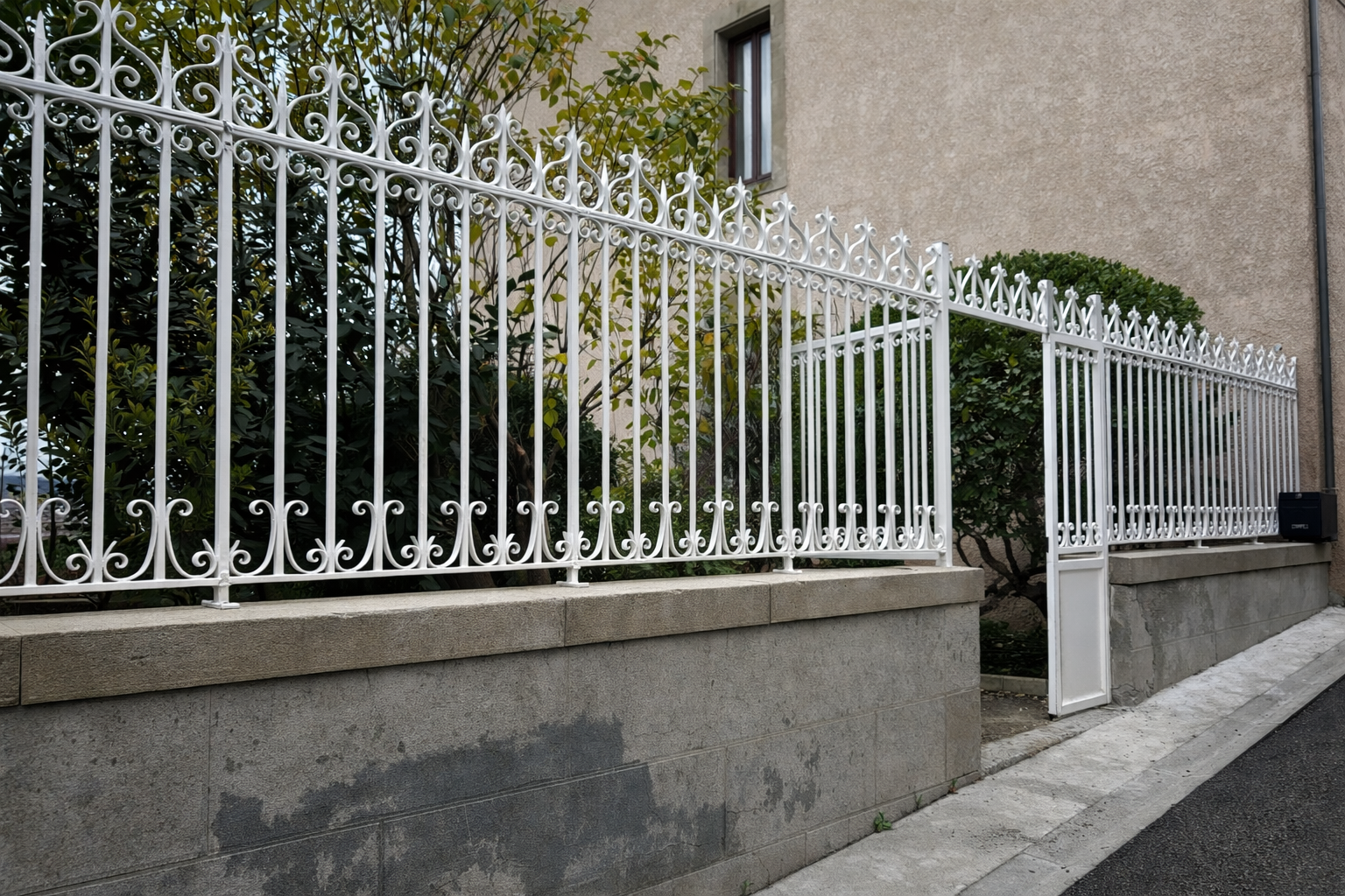 Clôture décorative en métal blanc sur un mur en béton, à côté d'un bâtiment et de verdure.