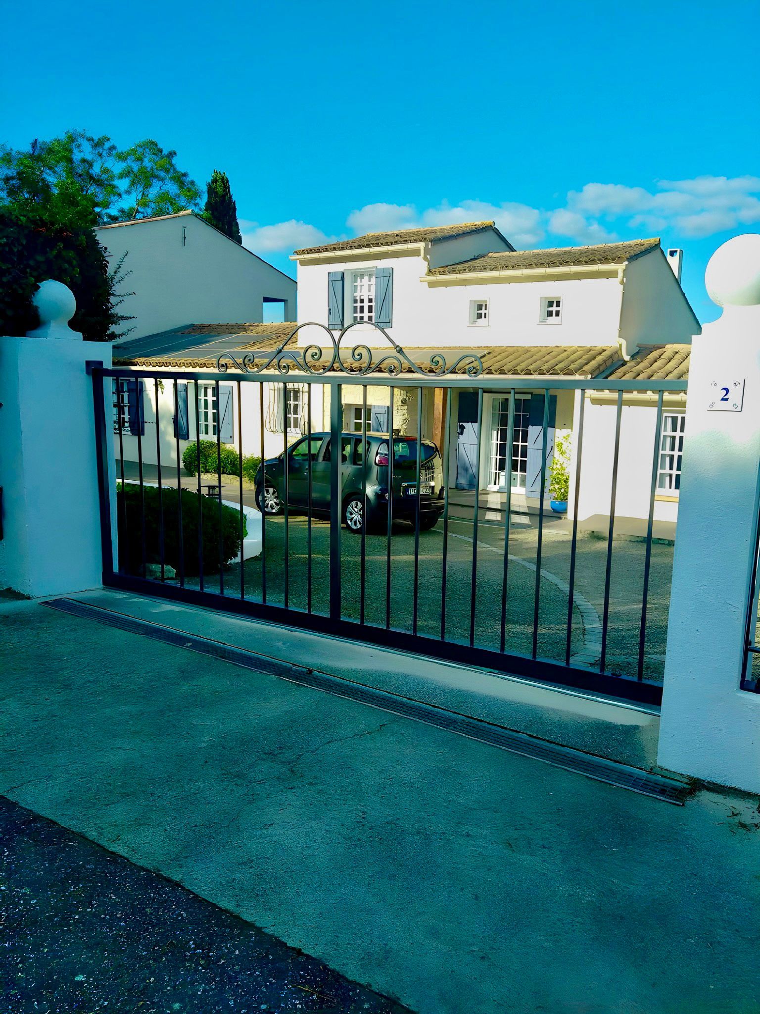 Portail en métal noir avec partie supérieure avec des volutes en fer forgé et barreaux verticaux, placé entre des piliers en béton.