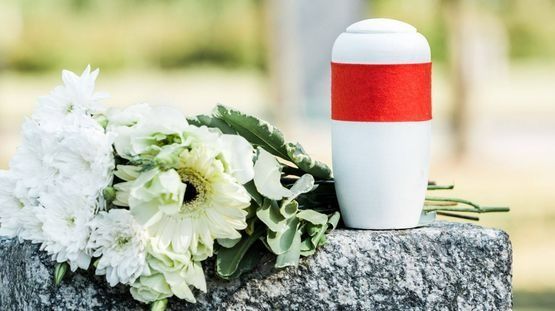 Una urna blanca con una cinta roja se encuentra encima de una tumba con flores.
