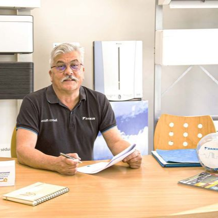 Un homme aux cheveux gris et à la moustache est assis à un bureau, des papiers à la main, dans une salle d'exposition climatisée.