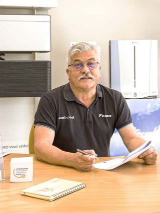 Un homme assis à un bureau, tenant des documents, regarde la caméra. Il porte une chemise noire. Scène en intérieur.