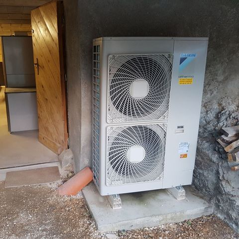 Unité de pompe à chaleur extérieure Daikin près d'une porte, avec un tuyau rouge sur une base en béton.