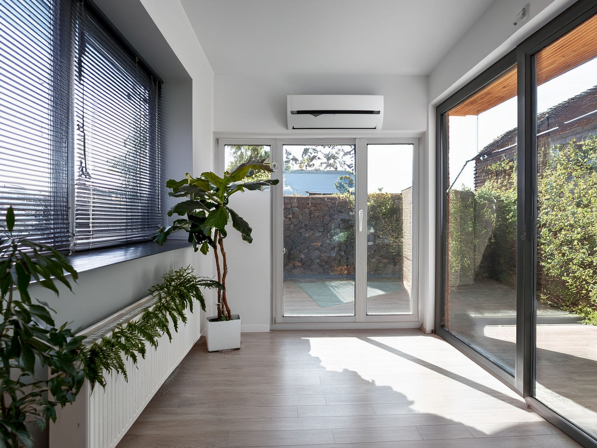 Véranda lumineuse avec grandes fenêtres, plantes, climatiseur et plancher en bois.