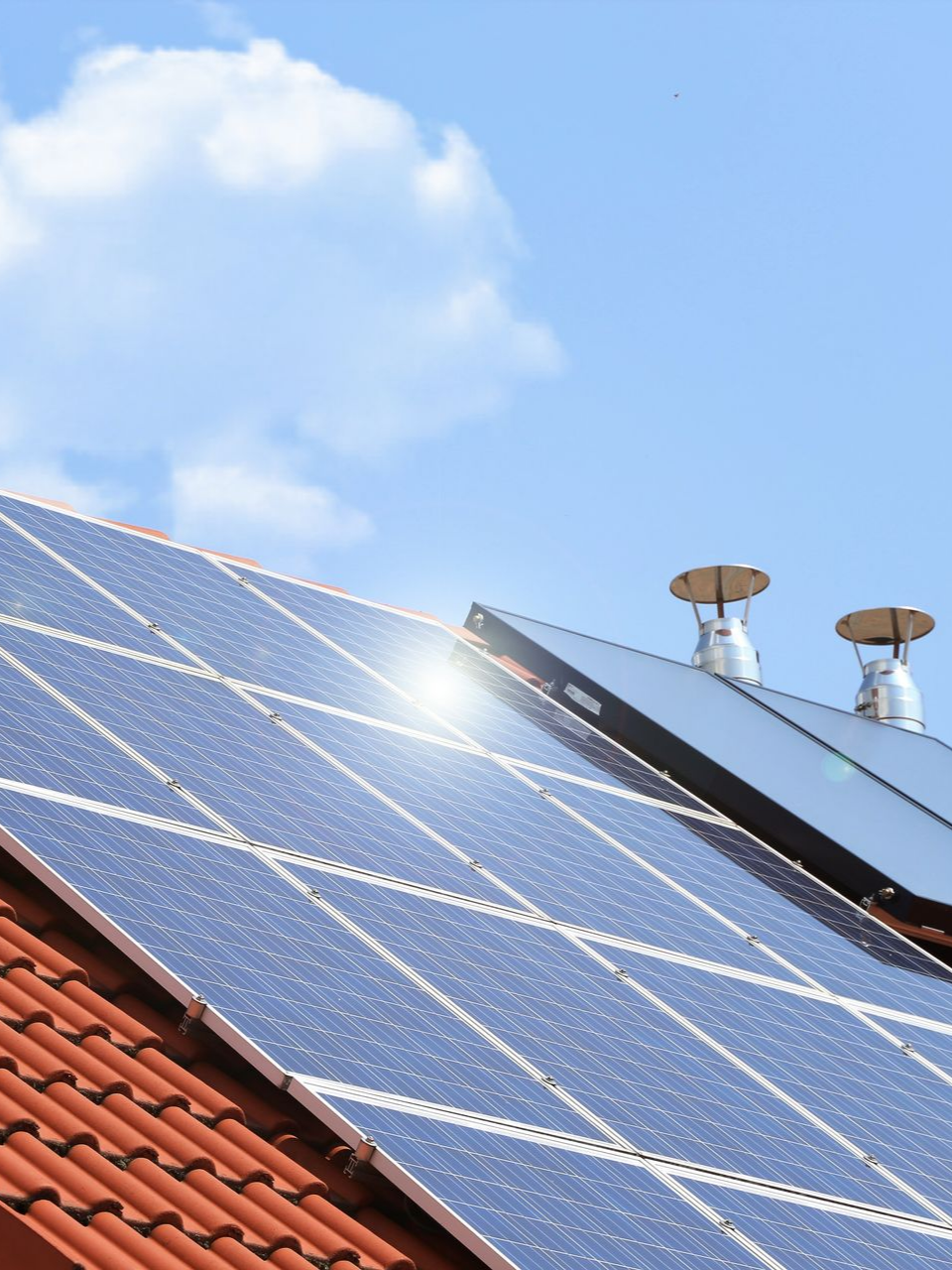 Des panneaux solaires sur un toit de tuiles rouges, avec deux cheminées, se détachant sur un ciel bleu.