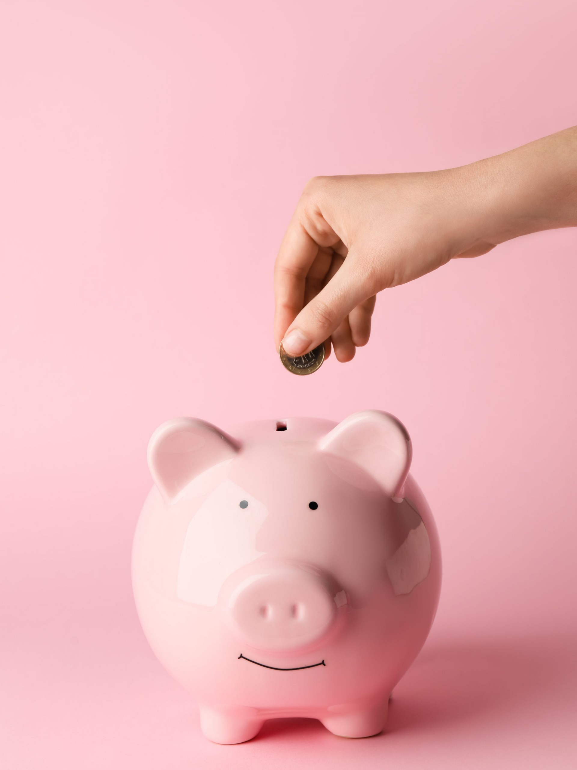 Une main déposant une pièce dans une tirelire rose sur fond rose.