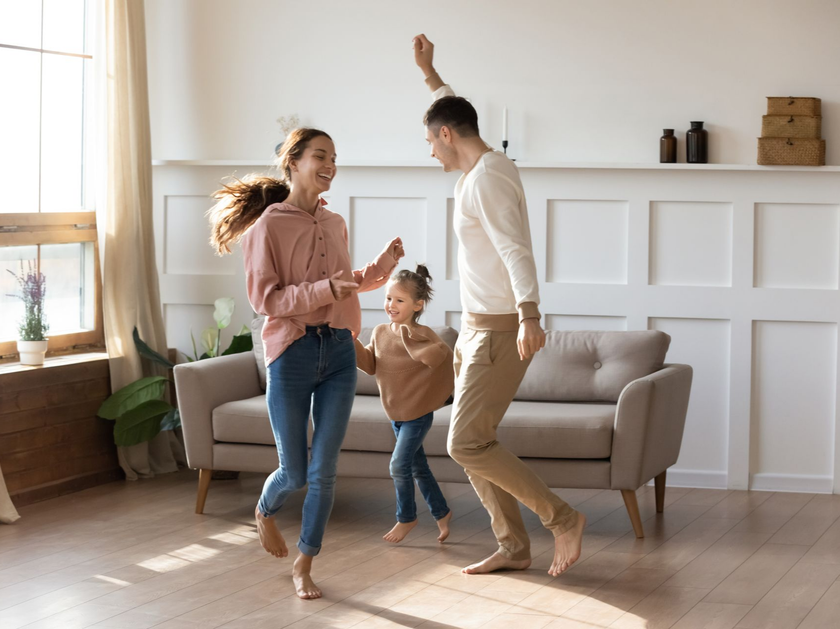 La famille danse pieds nus joyeusement dans le salon. La lumière du soleil entre par la fenêtre.