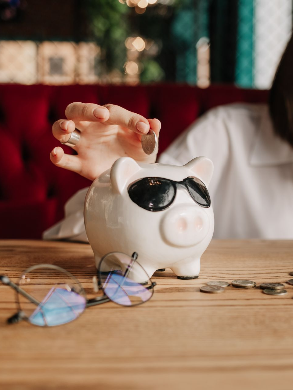 Une personne mettant une pièce dans une tirelire blanche, portant des lunettes de soleil ; des pièces et des lunettes sur une table.