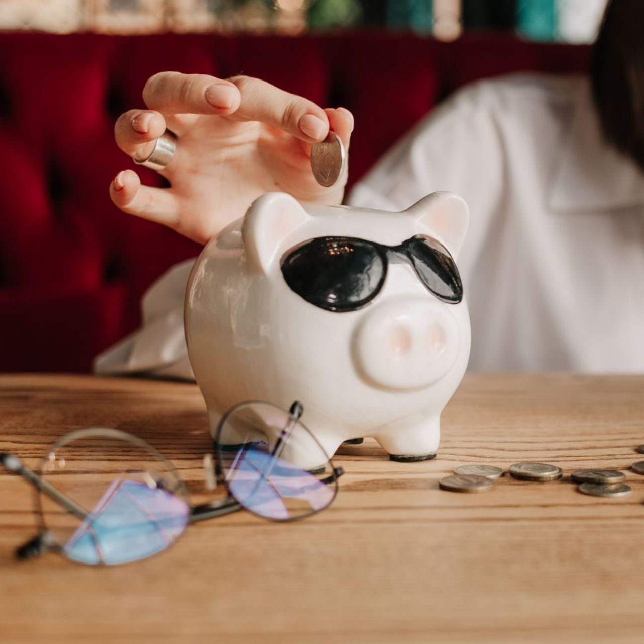 Une personne mettant une pièce dans une tirelire, portant des lunettes de soleil ; des pièces et des lunettes sur la table.