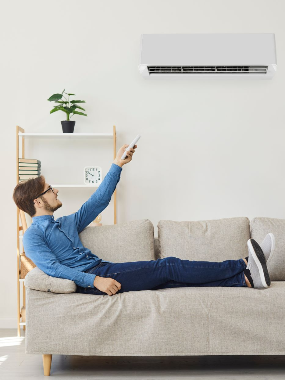 Un homme, assis sur un canapé, utilise la télécommande pour régler un climatiseur mural. Intérieur.