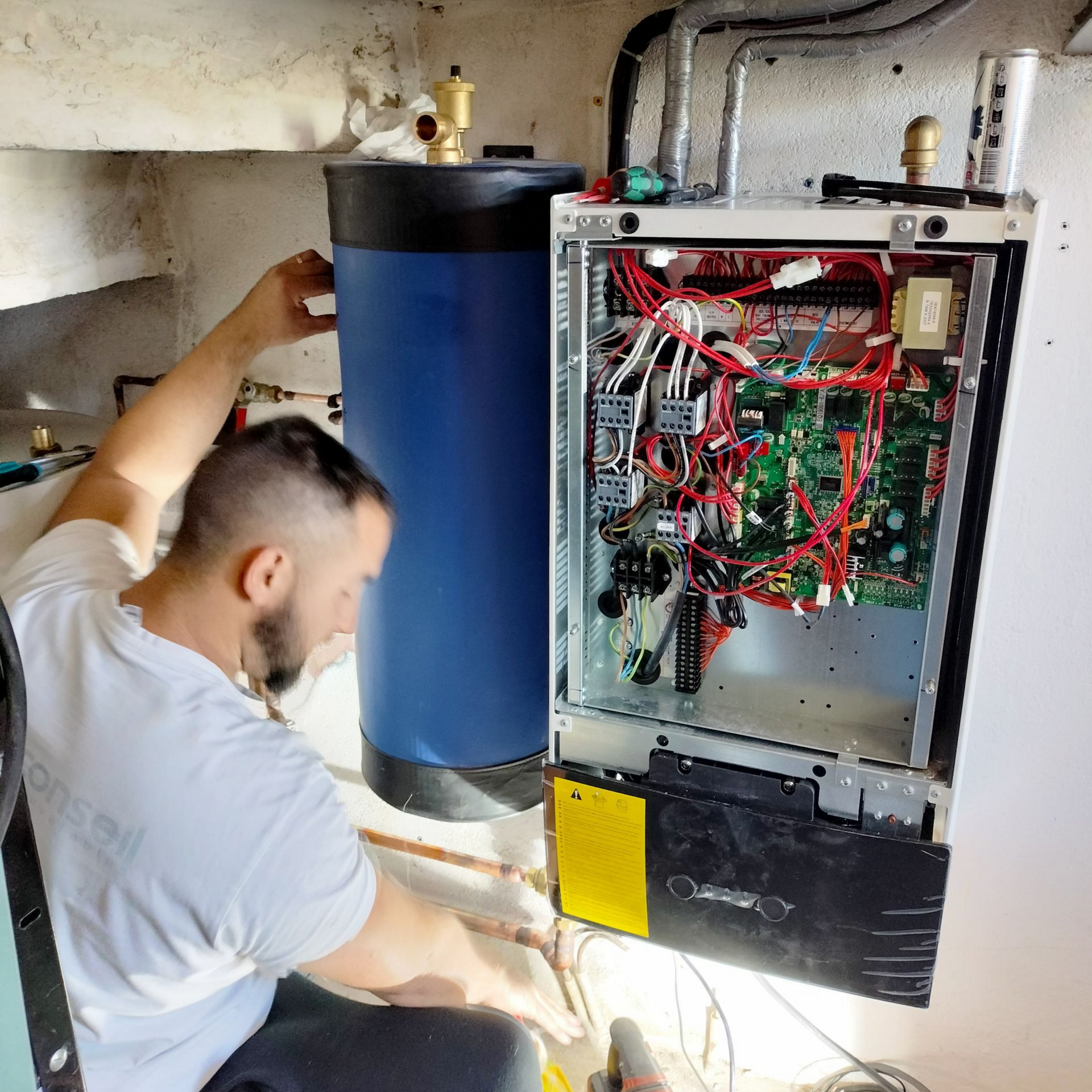 Une personne installe un chauffe-eau dans une buanderie. L'appareil est bleu et blanc, avec des fils électriques apparents.