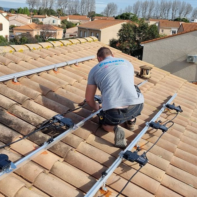 Un homme installe des panneaux solaires sur un toit de tuiles dans une zone résidentielle.