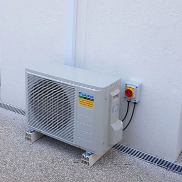 Un climatiseur extérieur Daikin avec un interrupteur marche/arrêt sur un mur blanc. Il repose sur des blocs de béton.