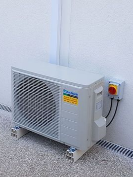 Unité de climatisation extérieure contre un mur blanc, avec un boîtier électrique adjacent.