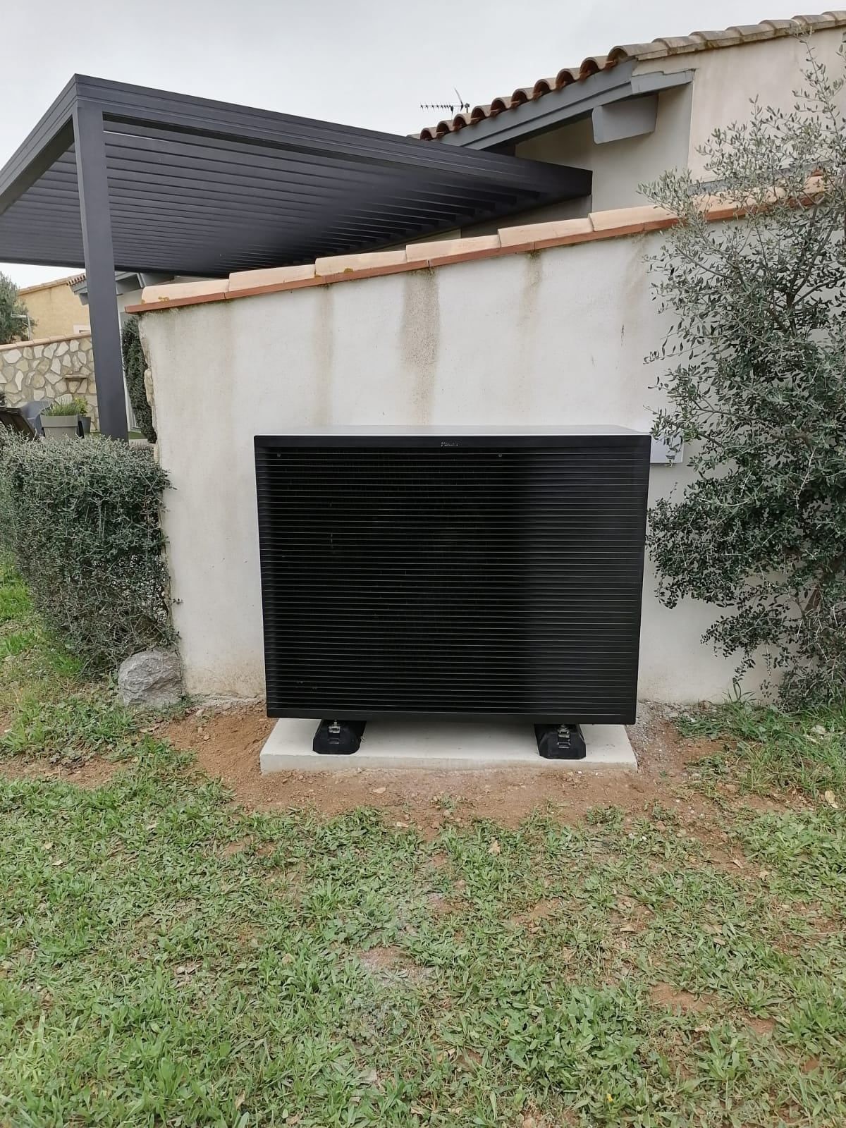 Pompe à chaleur rectangulaire noire contre un mur beige, sous une pergola, sur une base en béton, installée dans la pelouse.