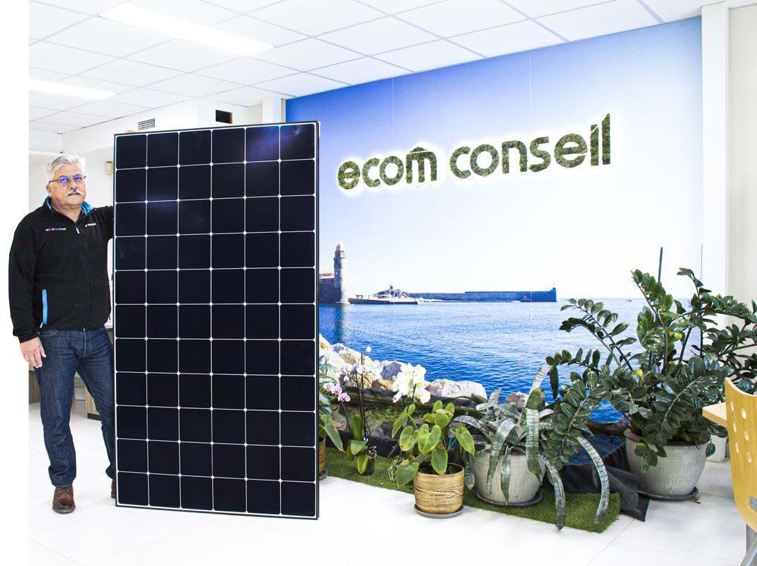 Un homme se tient à côté d'un grand panneau solaire à l'intérieur d'un bureau. Logo Ecom Conseil sur le mur.