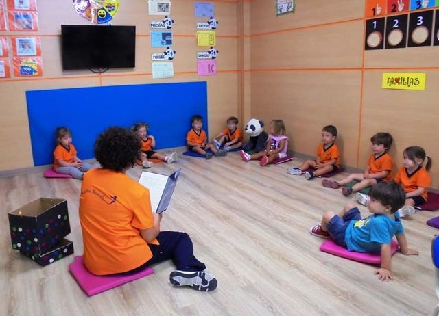 Una mujer con una camisa naranja está leyendo un libro a un grupo de niños.