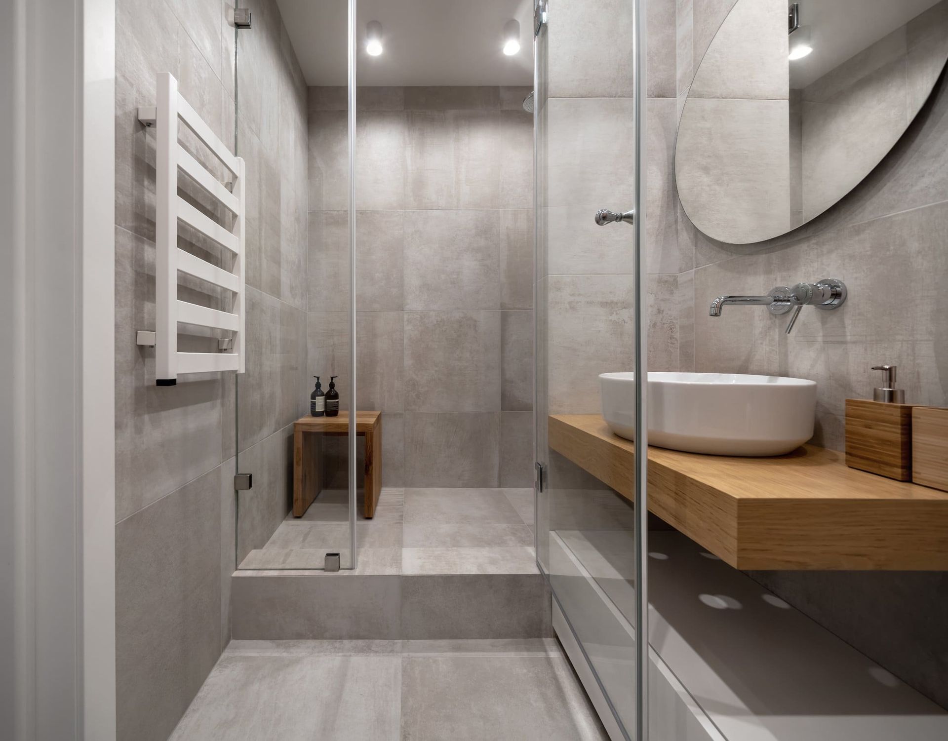 Baño moderno: azulejos grises, ducha de cristal, tocador flotante de madera con lavabo blanco, calentador de toallas y espejo ovalado.