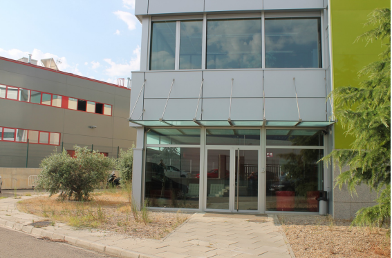 Entrada de edificio moderno con puertas de vidrio, acera y vegetación; otro edificio al fondo.