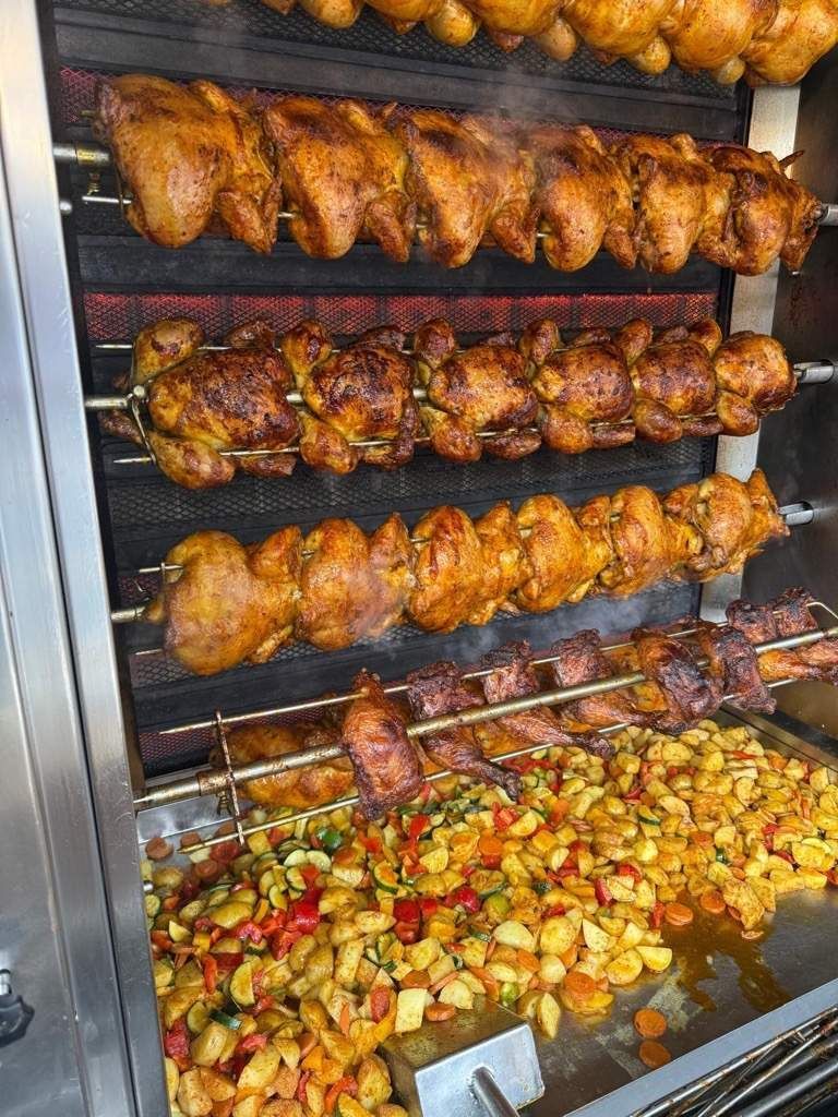 Brathähnchen braten am Spiess; darunter Kartoffeln und Gemüse.