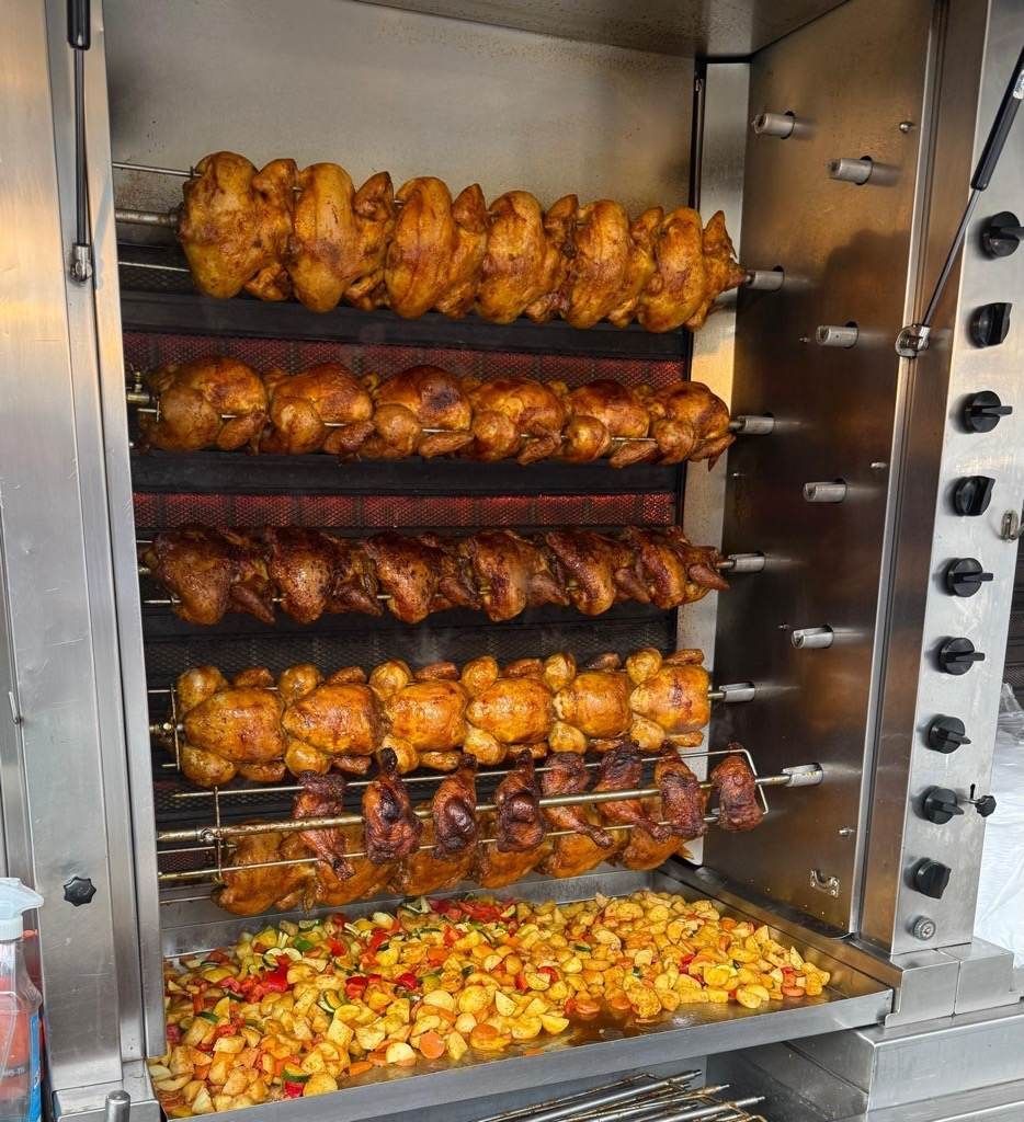 Ein Drehgrillofen mit mehreren Reihen gebratener Hähnchen, darunter Kartoffeln in einem Tablett.