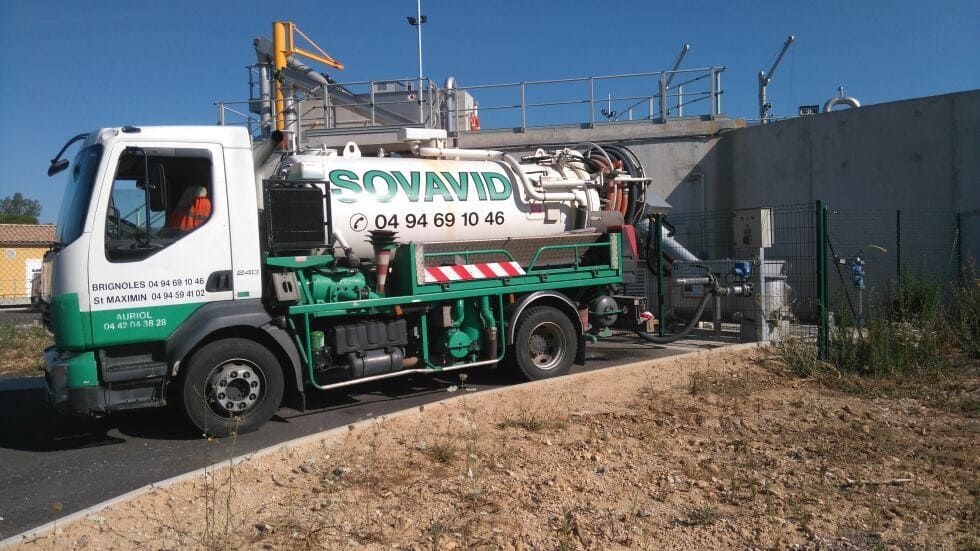 Un camion-citerne vert et blanc est garé à côté d'un mur en béton sur un terrain vague sous un ciel bleu dégagé.