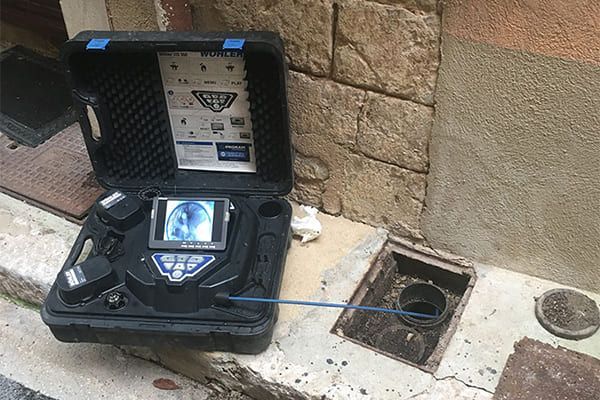 Boîtier noir ouvert avec un appareil bleu à côté d'un trou d'accès endommagé sur le trottoir et d'un mur de pierre.