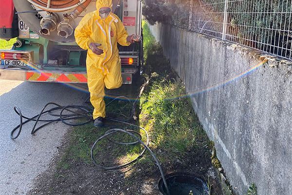 Un ouvrier en combinaison jaune utilise un tuyau d'arrosage pour nettoyer un fossé en bord de route, à côté d'un camion.