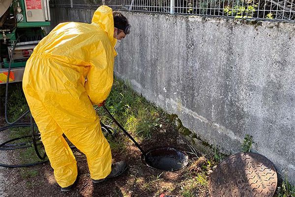Un ouvrier en combinaison de pluie jaune nettoie un caniveau à côté d'un mur en béton avec un tuyau d'arrosage.