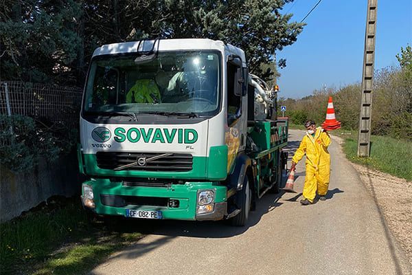 Un camion vert SOVAVID stationné en bord de route, avec deux ouvriers en combinaisons de protection jaunes et des cônes orange à proximité.